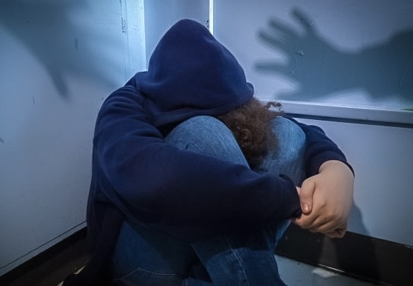 A student sits curled up in a corner, hoodie over their head with their face in their. Shadowed hands are reaching toward them.